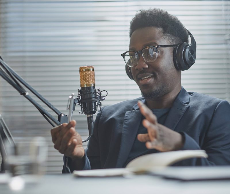 Medium close up shot of young Black man wearing headphones and eyeglasses speaking and adjusting microphone stand during live podcast