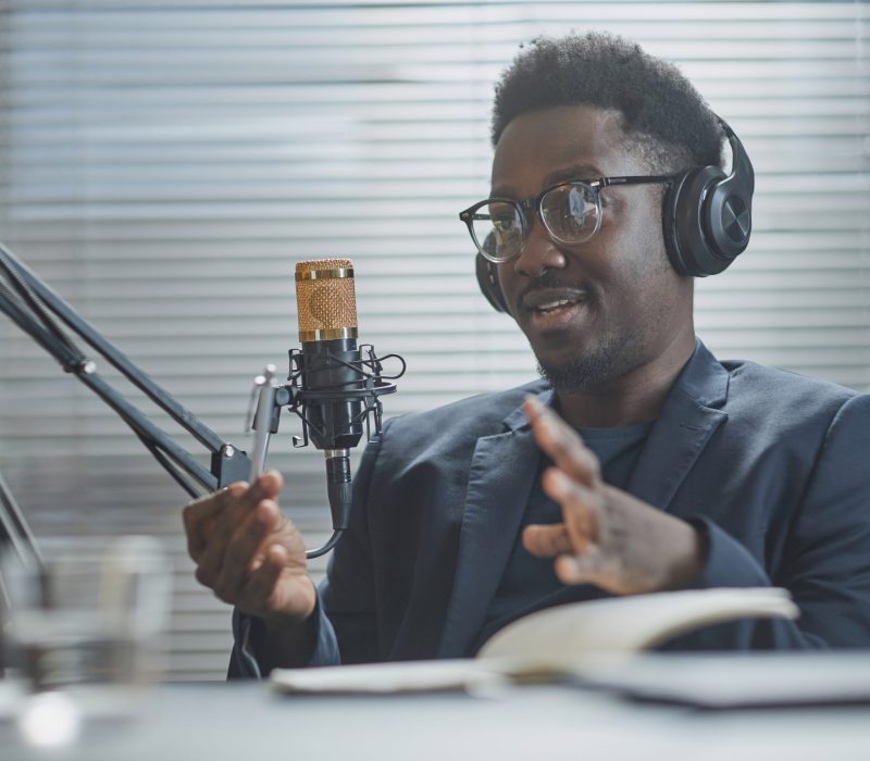 Medium close up shot of young Black man wearing headphones and eyeglasses speaking and adjusting microphone stand during live podcast